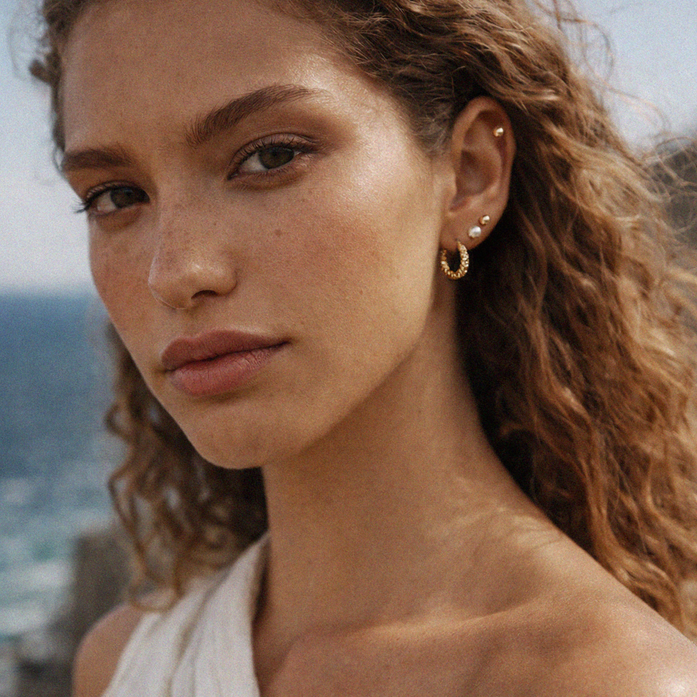 Bizou SF253E006GL – Woman wearing gold stainless steel and pearl earring set by the sea, ear stack styling, Mediterranean lifestyle, Bizou jewelry