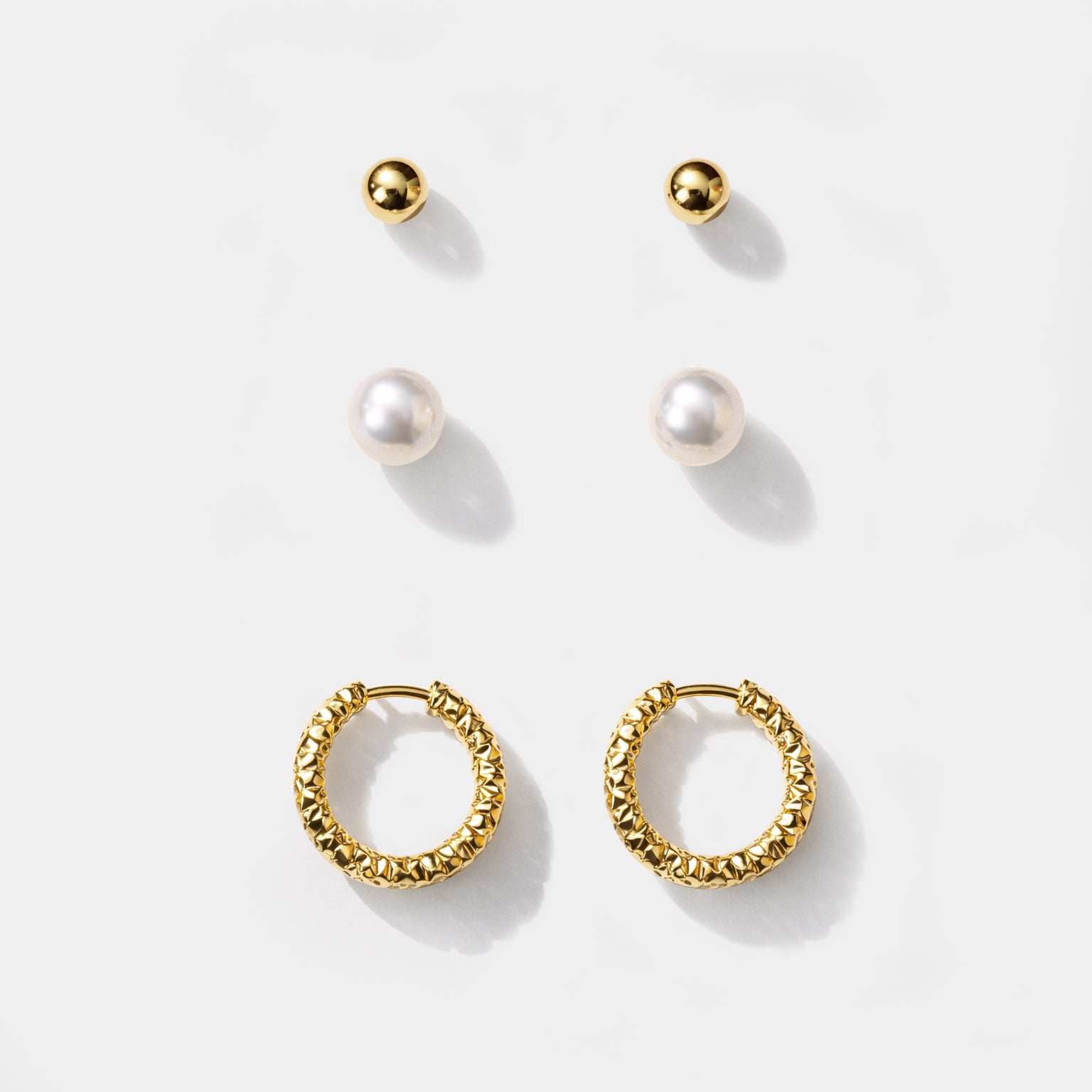 Bizou SF253E006GL – Gold stainless steel earring set of 3 pairs, ball studs, pearl studs and textured huggie hoops, flat lay view